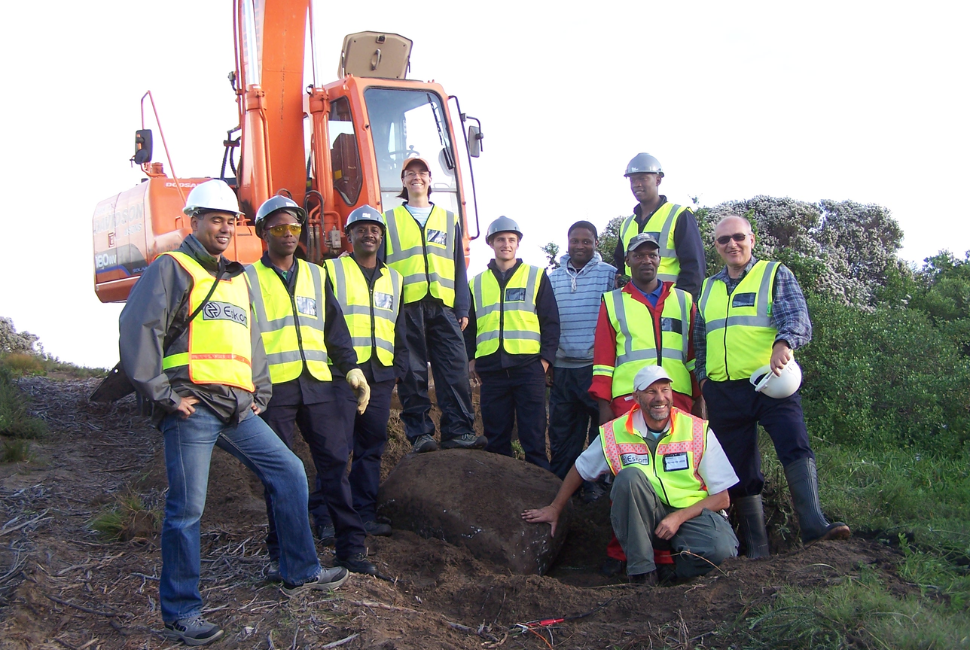 A group of people wearing hi-vis safety vests and hard hats pose for a photo in front of an excavator during a seismic hazard assessment.