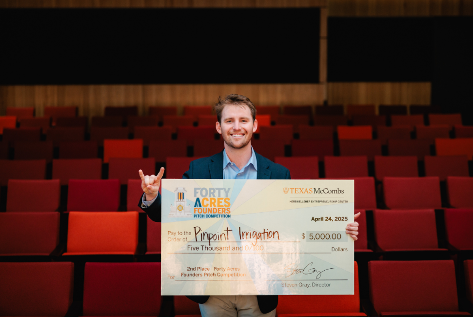 A man smiles and holds up the Hook 'Em, Horns hand sign while posing for a photo with a large check in an empty auditorium.