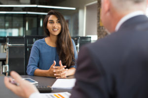 A young businesswoman sitting at table, talking to senior male manager and smiling in office.