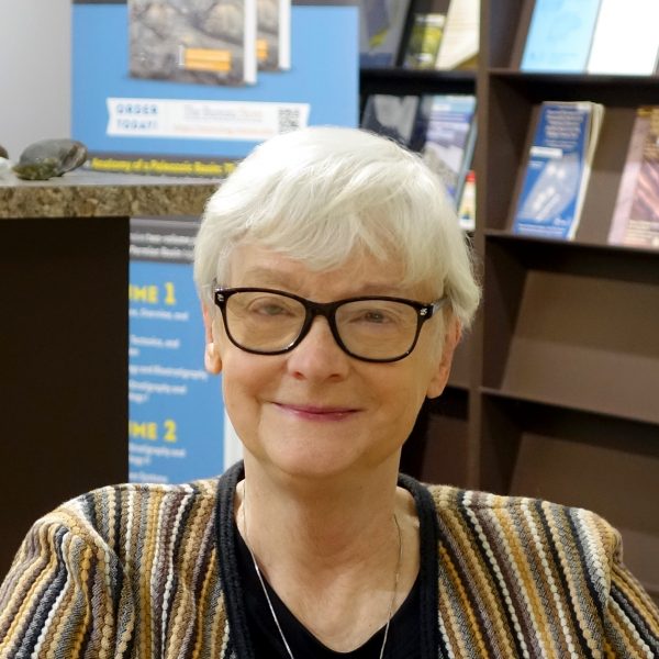 A woman wearing glasses sits in front of some shelves with pamphlets. She has short white hair and wears a striped jacket.