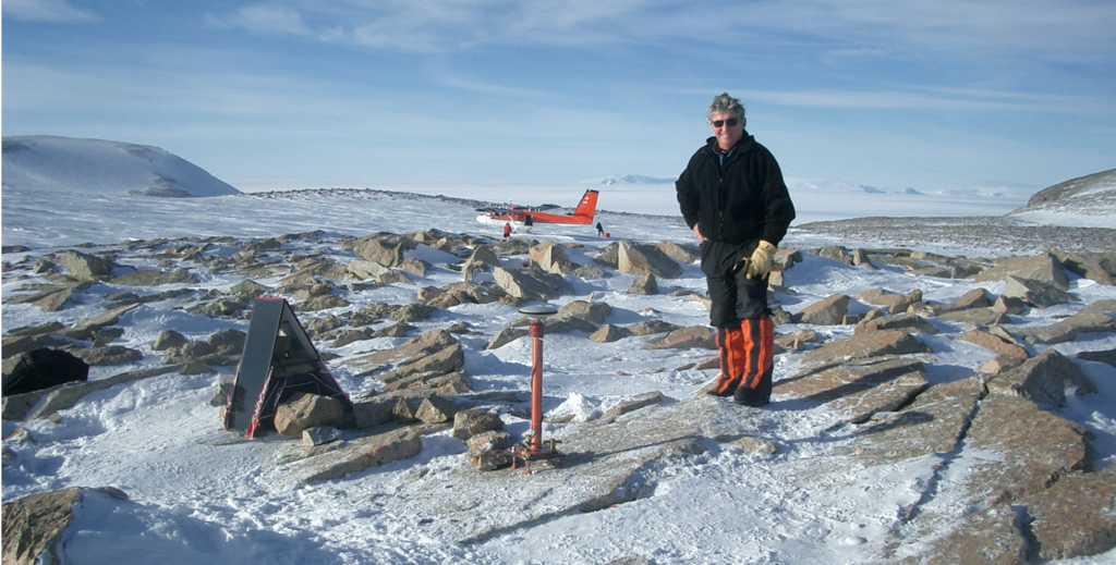 A picture showing Ian Dalziel standing among snow and rocks with a deployed sensor ahead of him. In the background is a landed Twin Otter aircraft.