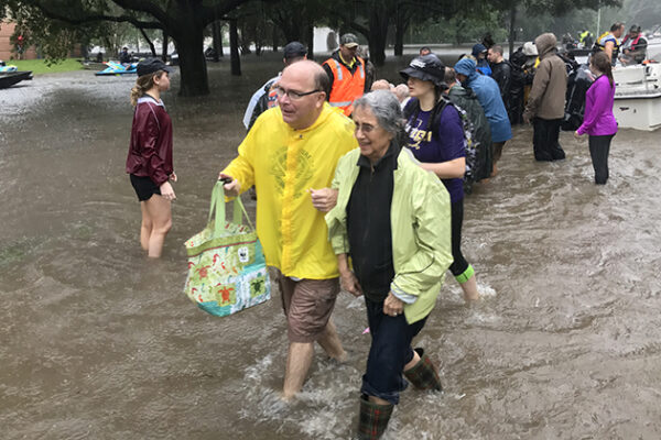A street flooded with brown water. A man in a yellow rain slicker walks arm in arm with a woman in rain boots in the ankle deep water.
