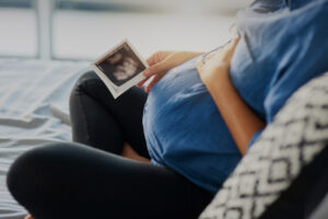 Image shows seated pregnant person from the shoulders down, wearing a blue shirt and holding an ultrasound photo of an infant in hand.