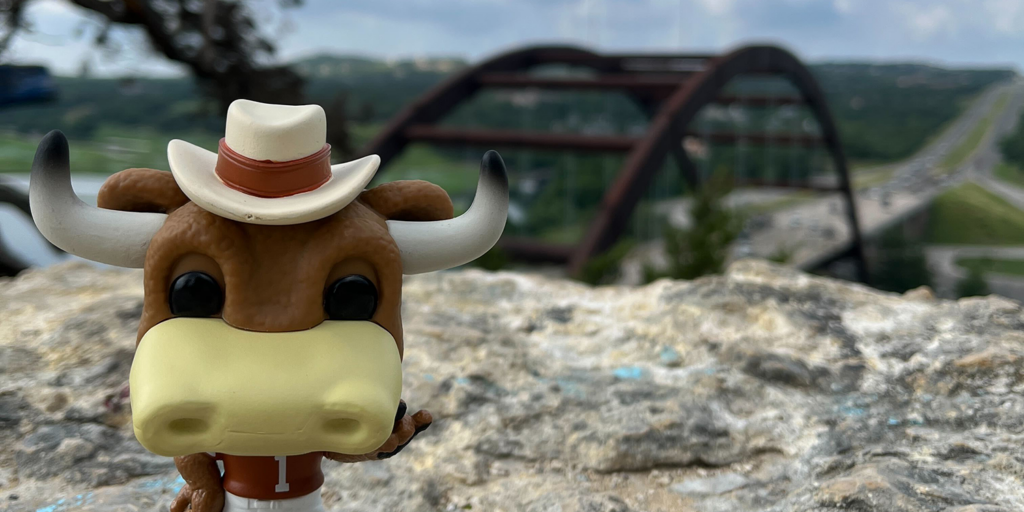 A Hook Em figurine stands on a cliff above Lake Austin with the Pennybacker Bridge in the background.