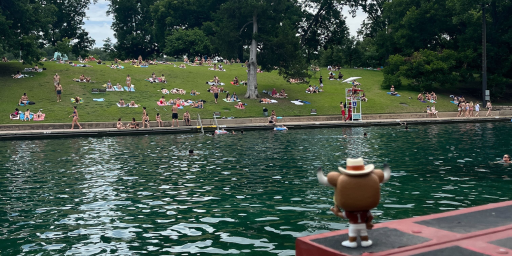 With its back to the camera, a figurine of Hook Em stands on a platform next to Barton Springs. A large number of people are gathered and relaxing on the grassy slope in the background. 