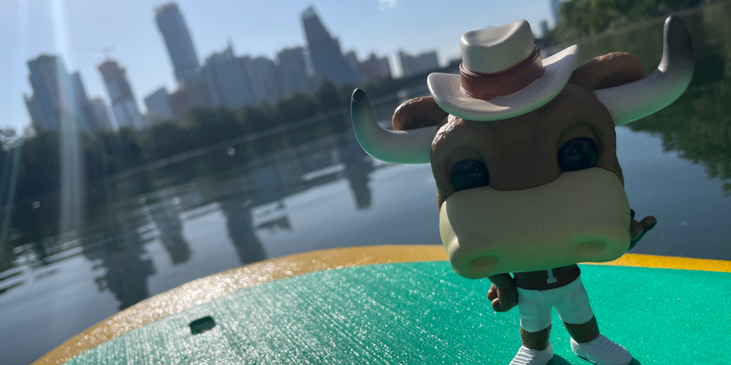 A Hook Em figurine stands on a paddleboard in the middle of Lady Bird Lake with the downtown Austin skyline in the background. 