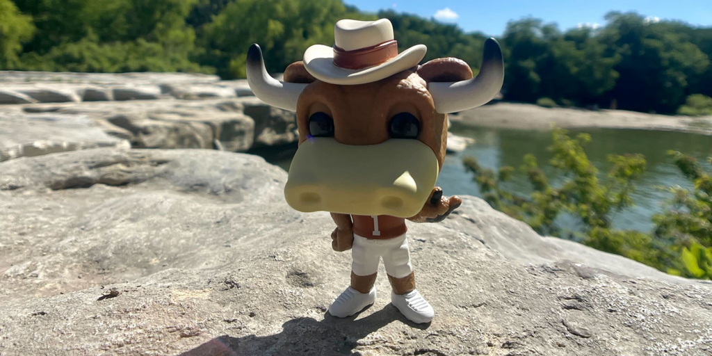 A Hook Em figurine stands on a rock with a natural pool of water in the background. 