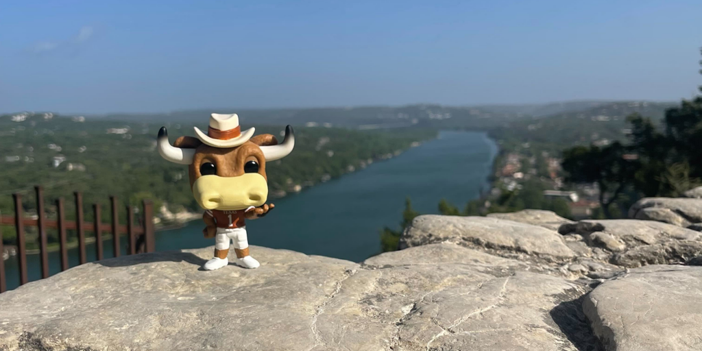 A figurine of Hook Em stands atop Mount Bonnell with Lake Austin and the sky in the background.