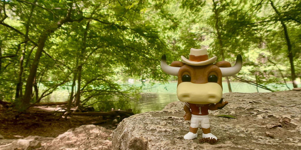 Lush green vegetation and cool, blue water in the background with a figurine of Hook Em standing in the foreground. 