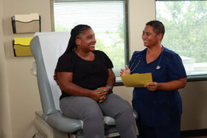 A Black woman, left, sits on an exam table while a Black woman doctor stands on the right as the two converse.