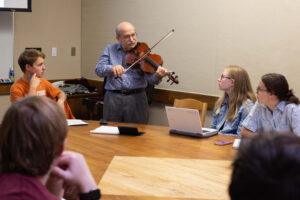 Michael Domjan holds a violin in front of students sitting around a table