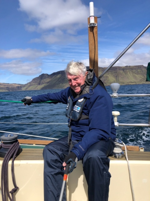 Photo of Dalziel sitting on the gunwale (side) of a boat at sea. The rocky green shore is about 2-3km away.