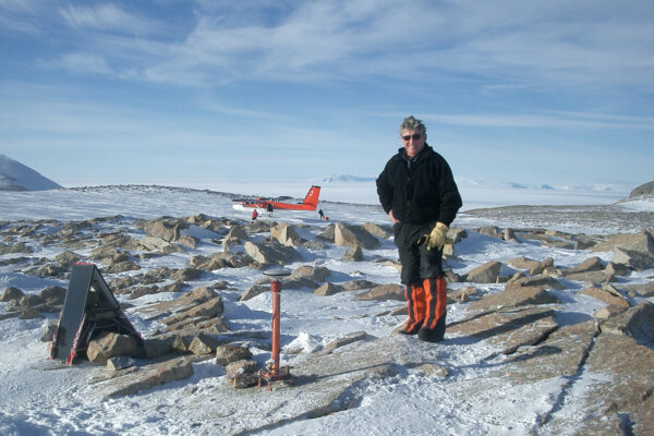 Alt: Photo of Dalziel in Antarctica. The landscape is rocky and ice covered. He is standing next to a waist-high GPS antenna. Parked behind him is a red and white Twin Otter aircraft.