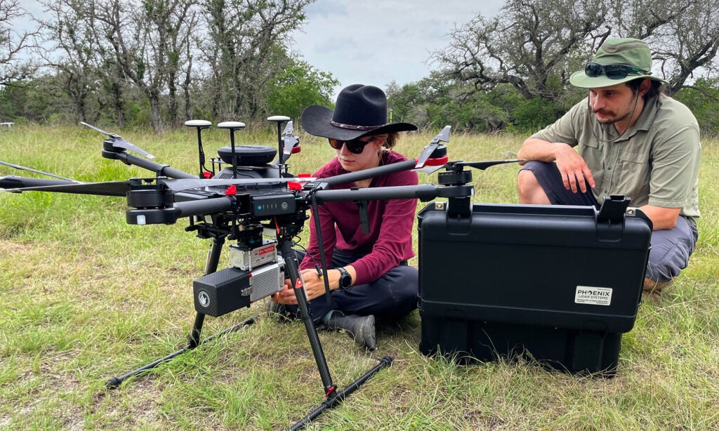 A drone like device about two feet tall is prepared by two researchers in an outdoor grassy and tree-ringed area