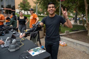 researcher standing at a table with rehabilitative robotic sleeve on right arm and doing hook 'em hand signal in left hand