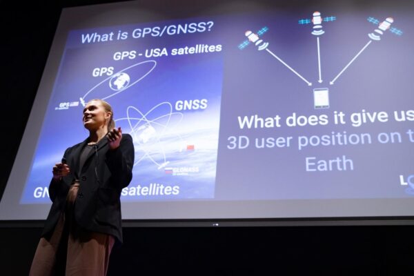 person stands in front of a screen with gps words, information, presenting to crowd