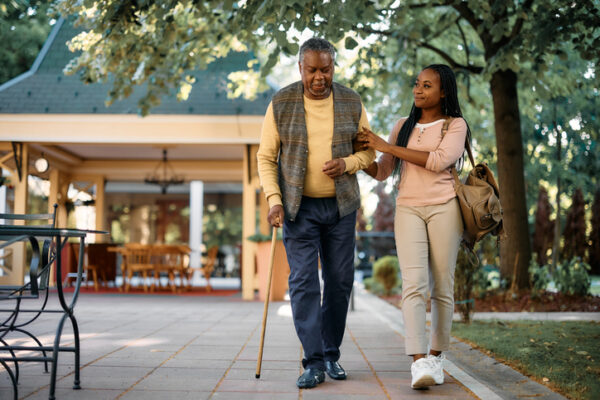Black senior man with walking cane and his daughter taking a walk through the park of a nursing home.