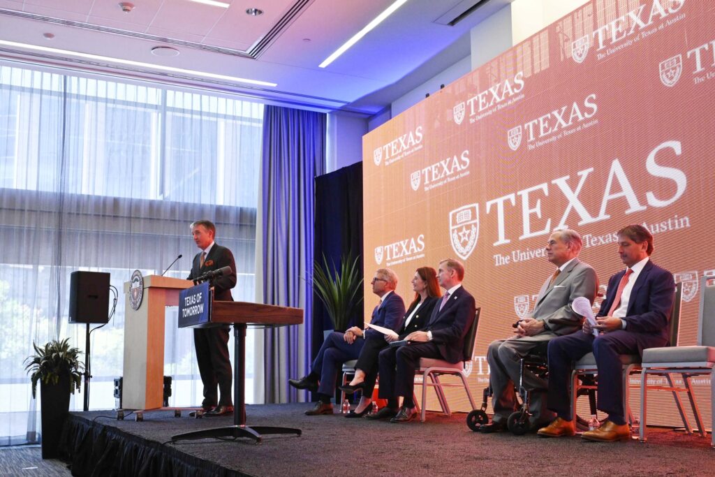 university leadership sits on a stage with president hartzell at the podium with a burnt orange backdrop and the Texas wordmark
