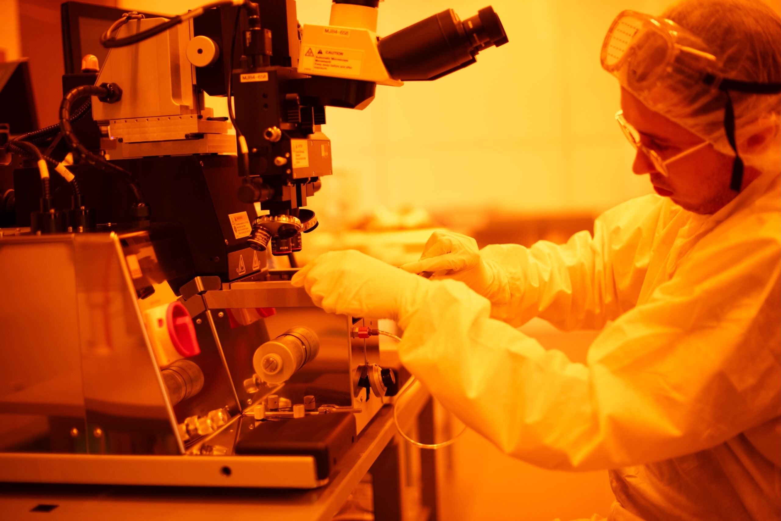 A student works on a microscope in a cleanroom with amber lighting.