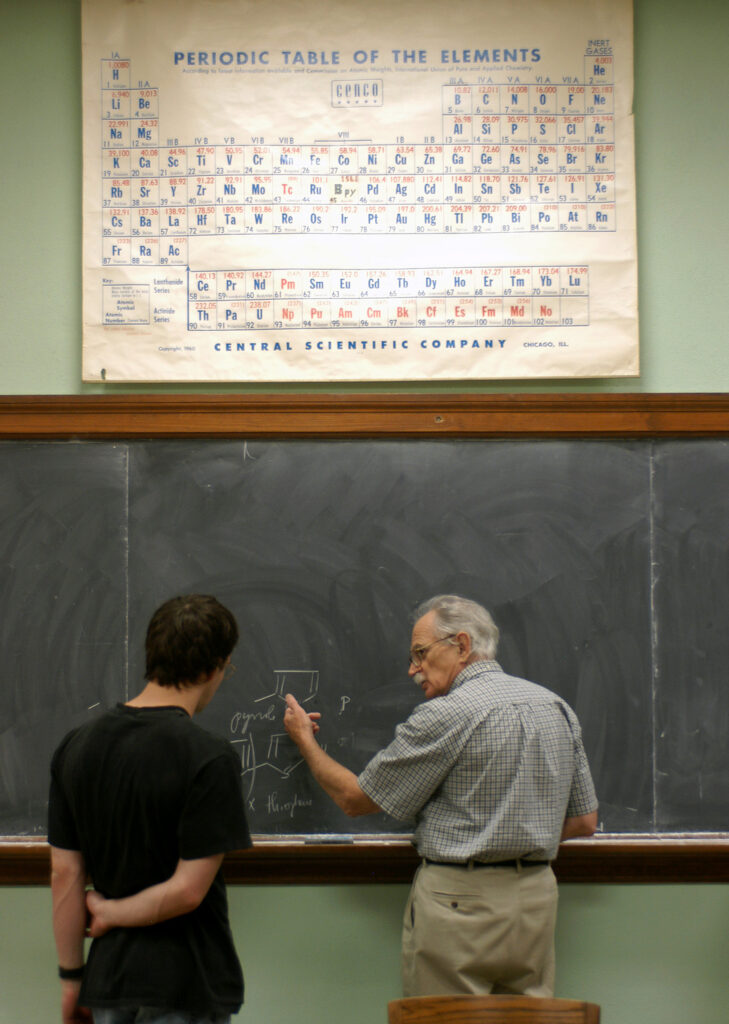 A scientist and a student stand at a chalkboard working through a chemistry problem