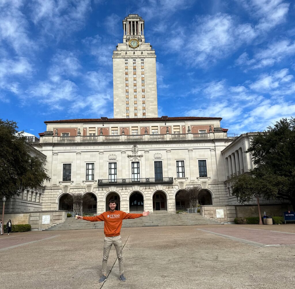 A man stands with his arms outstretched in front of the UT Tower's, wearing a spirit shirt