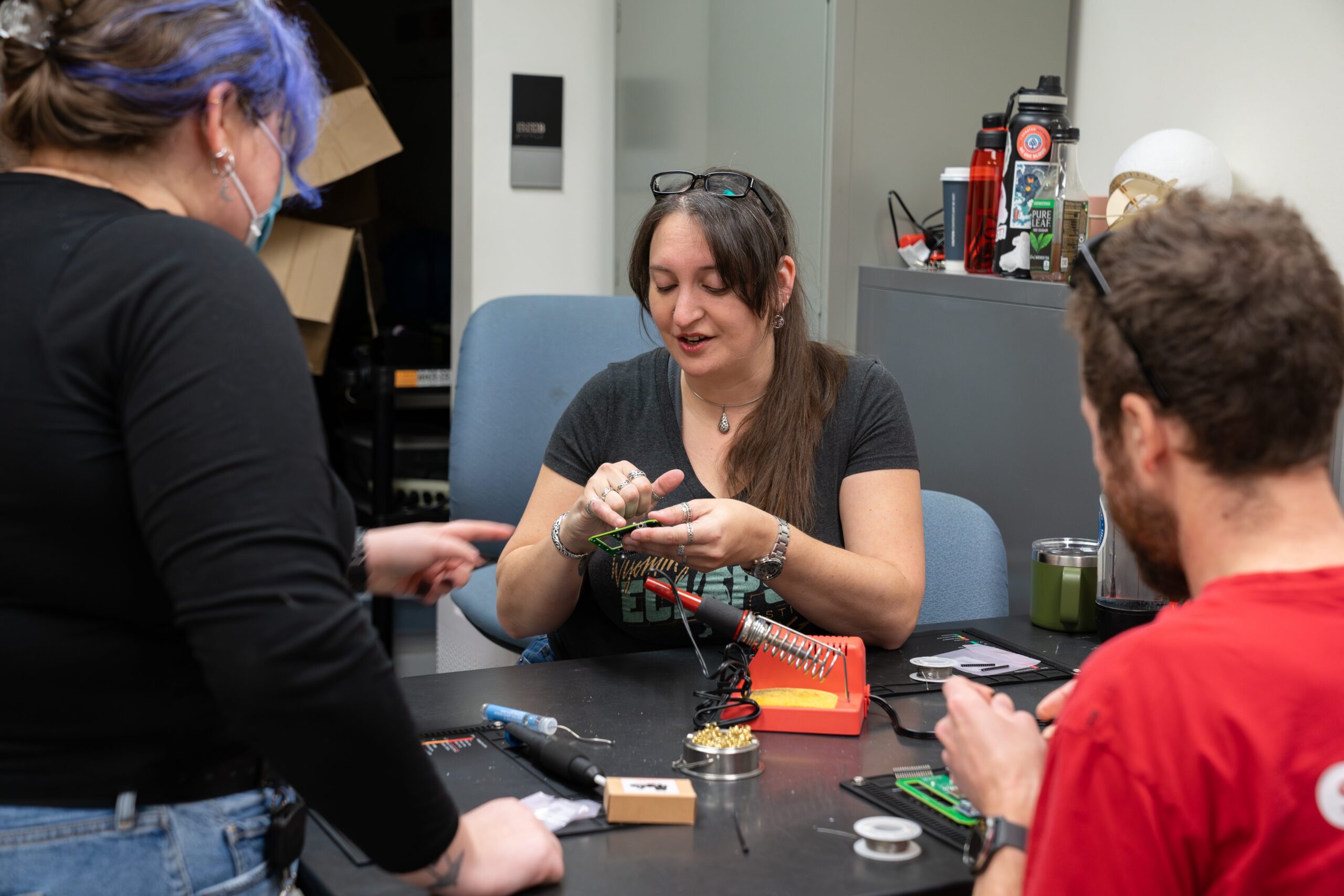 person pointing to a device while talking to another person with purple hair and person with red t-shirt sitting in foreground