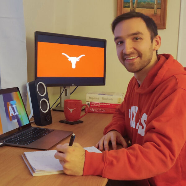 A man in a UT sweatshirt takes notes at his desk and smiles as the Longhorn silhouette and the words AI appear on screens nearby