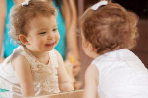 A toddler girl with brown curly hair looks in the mirror and smiles at her reflection.