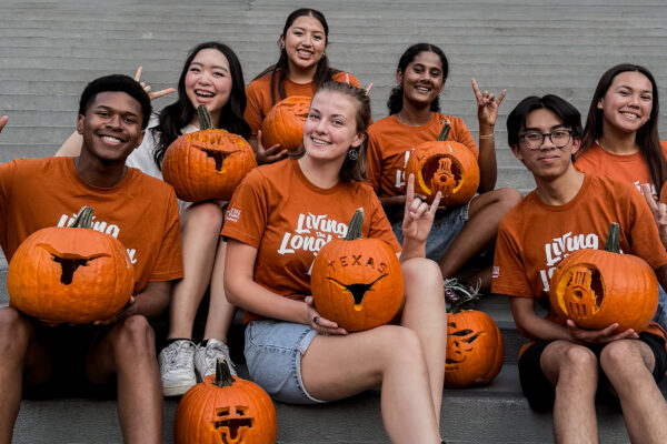 A group of seven U.T. Austin students pose together while holding jack-o'-lanterns carved with U.T. themed designs like the Longhorn logo and the U.T. Tower.