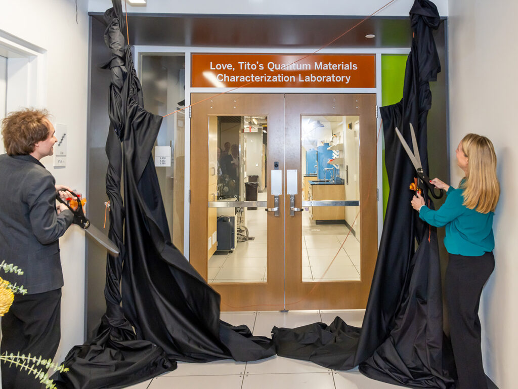 Two people with large scissors cut the cords on a pair of black drapes, revealing a new sign over the entrance to a lab. The sign reads: 