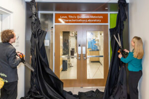 Two people with large scissors cut the cords on a pair of black drapes, revealing a new sign over the entrance to a lab. The sign reads: 