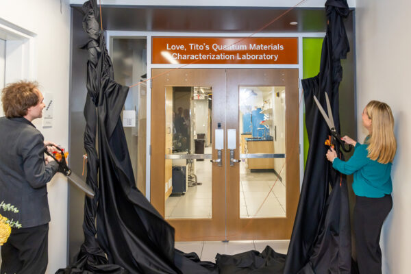 Two people with large scissors cut the cords on a pair of black drapes, revealing a new sign over the entrance to a lab. The sign reads: 