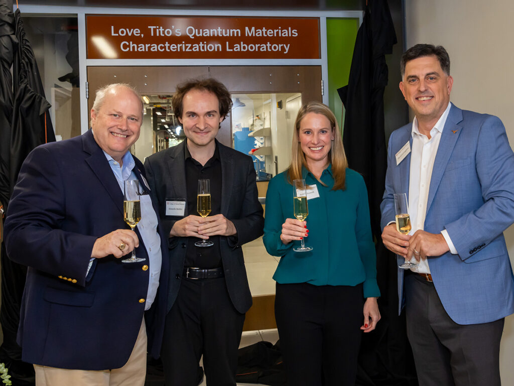 Four people hole champagne glasses and smile for the camera in front of the newly named Love, Tito's Quantum Materials Characterization Laboratory.