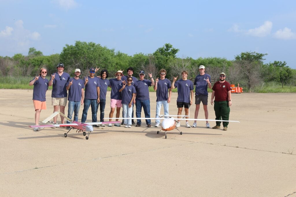 Multiple students with blue t-shirts stand on concrete near two drones
