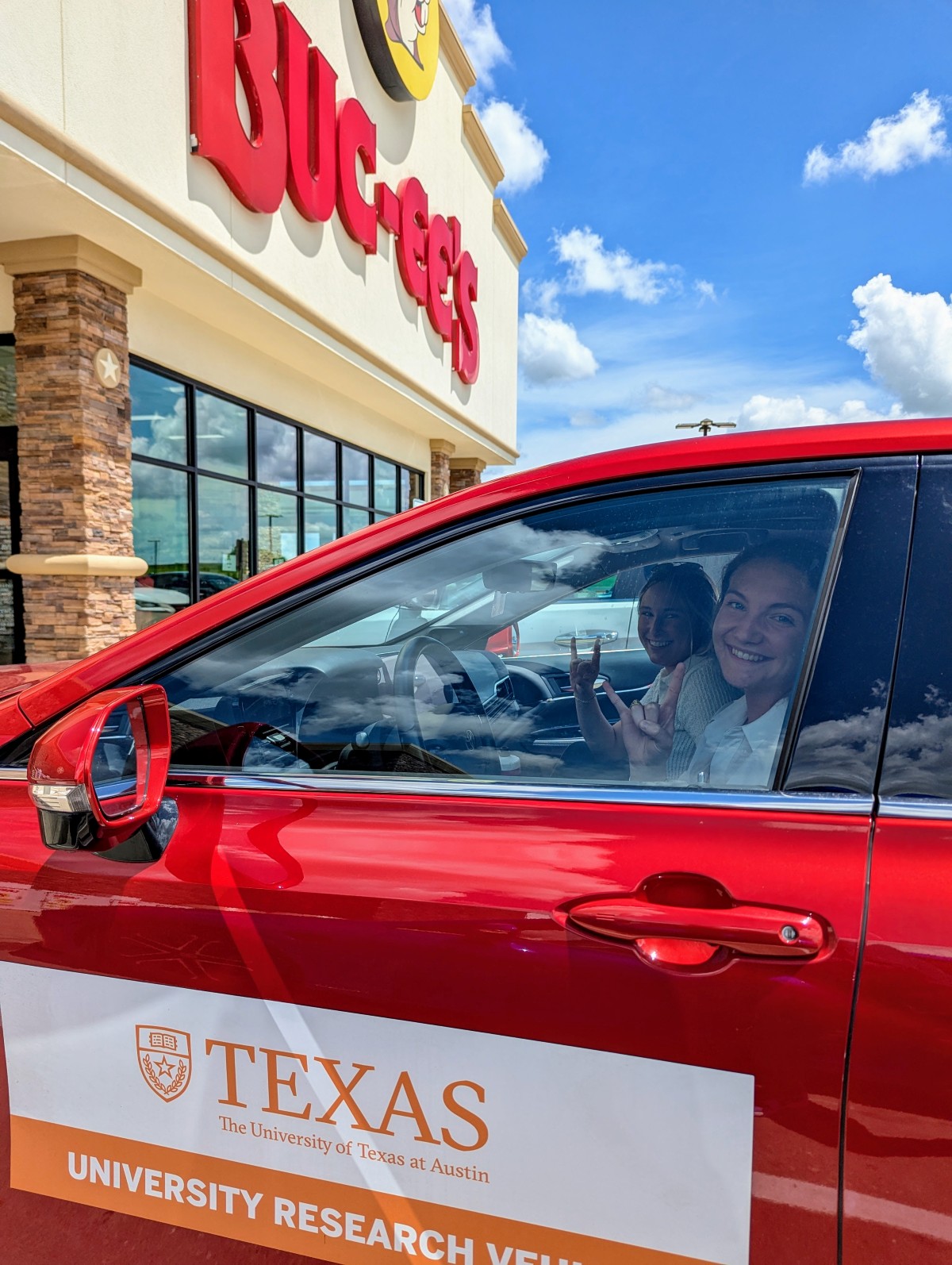 Two women sit in red car in front of Buc-ee's