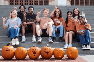 A group of students poses for a photo in front of six carved pumpkins.