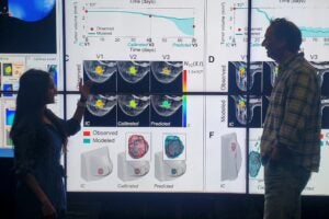 Two people stand in front of a graphic on a screen.