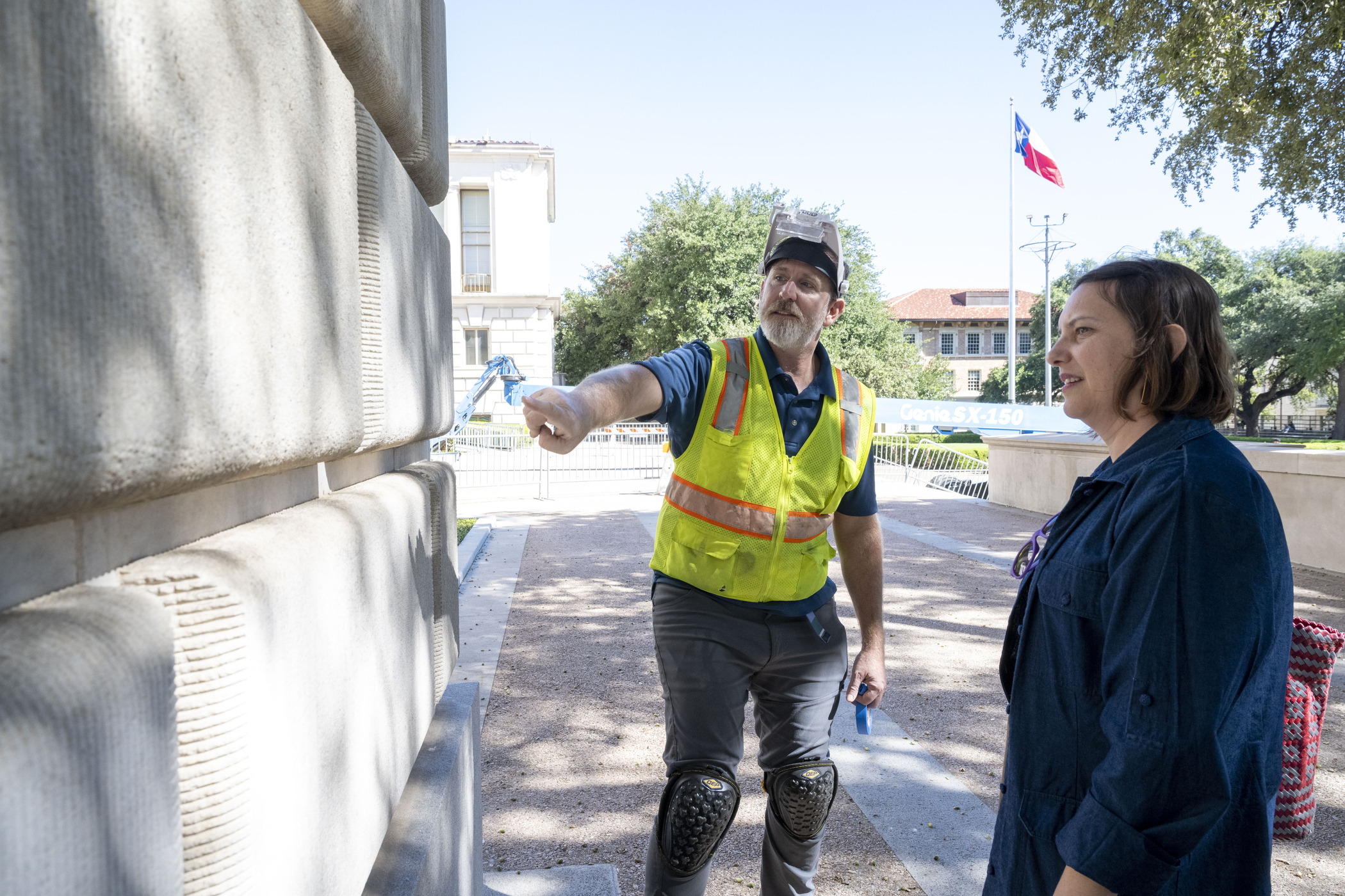 man in yellow construction vest points to stone siding of building as woman in blue dress listens