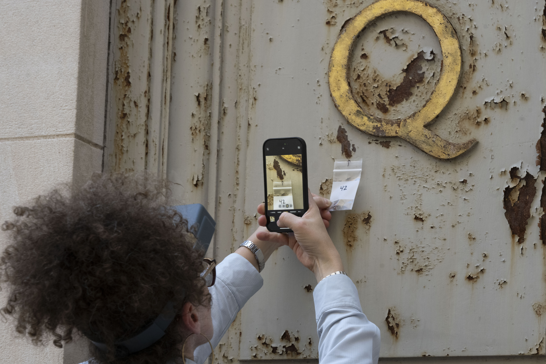 woman takes photo on phone of number on slip of paper she is holding up beneath the gilded letter Q