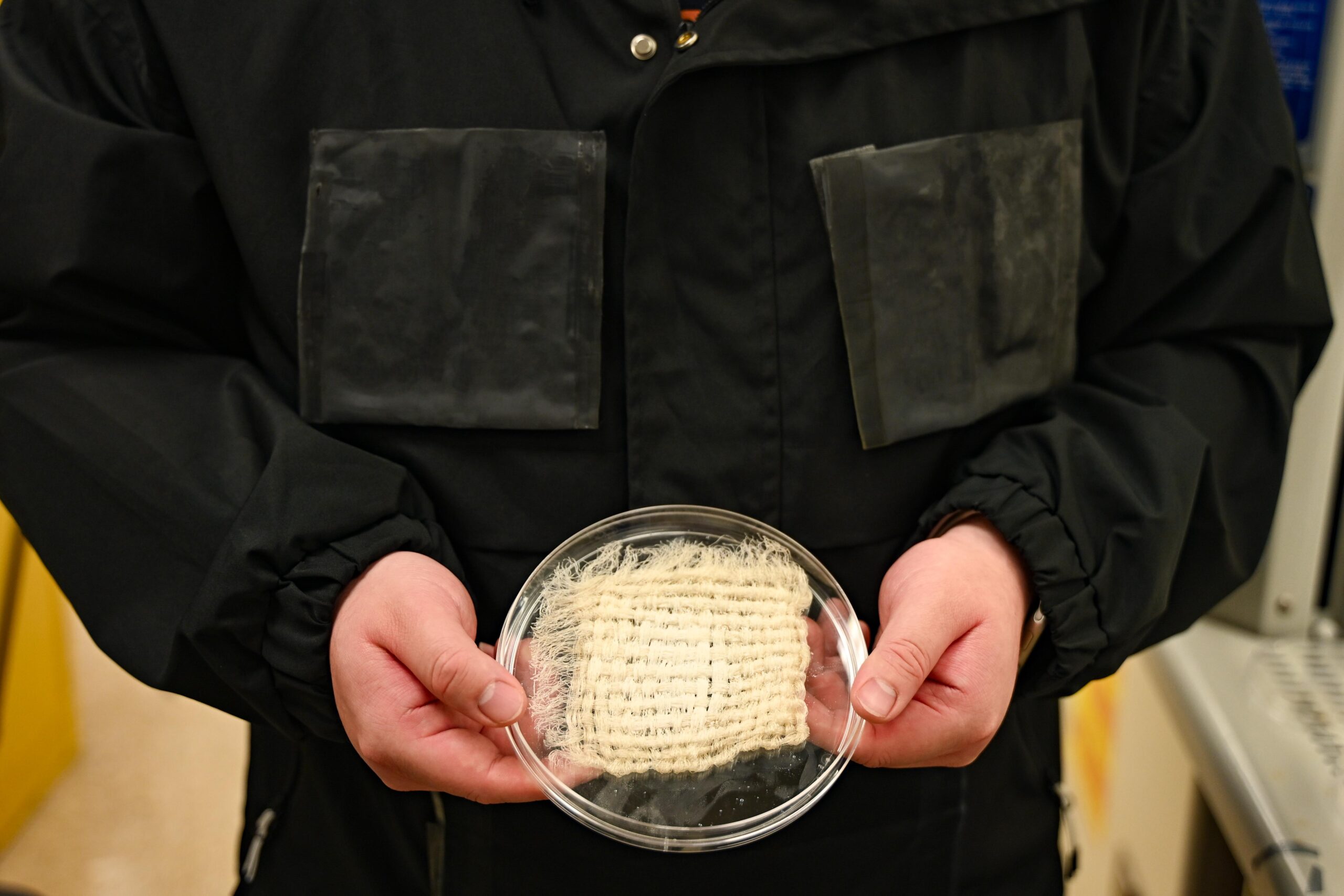 A researcher in a black jacket with pockets holds a beige-colored textile.