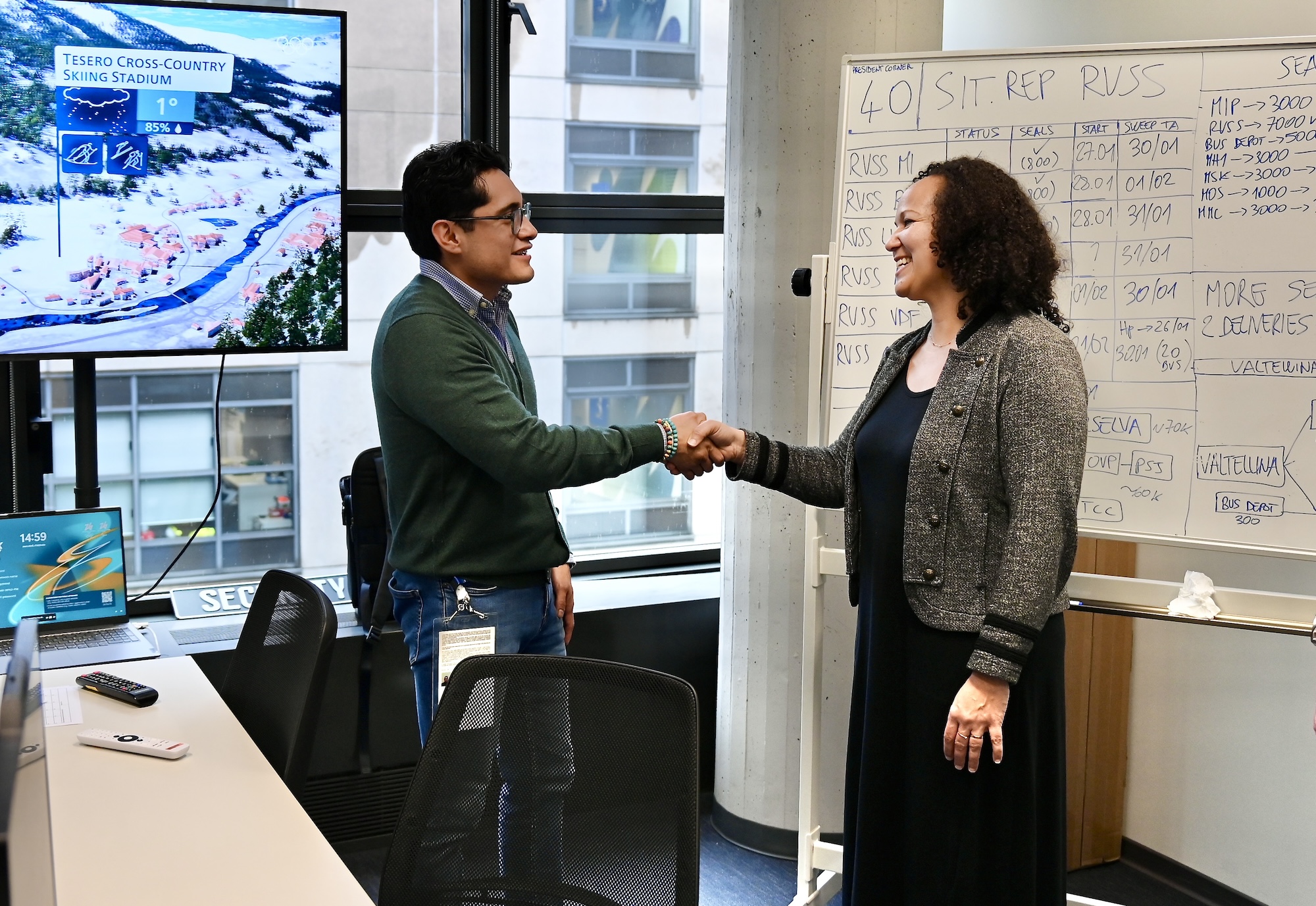 A man and a woman shake hands in a conference room. In the background, a TV screen and a whiteboard.