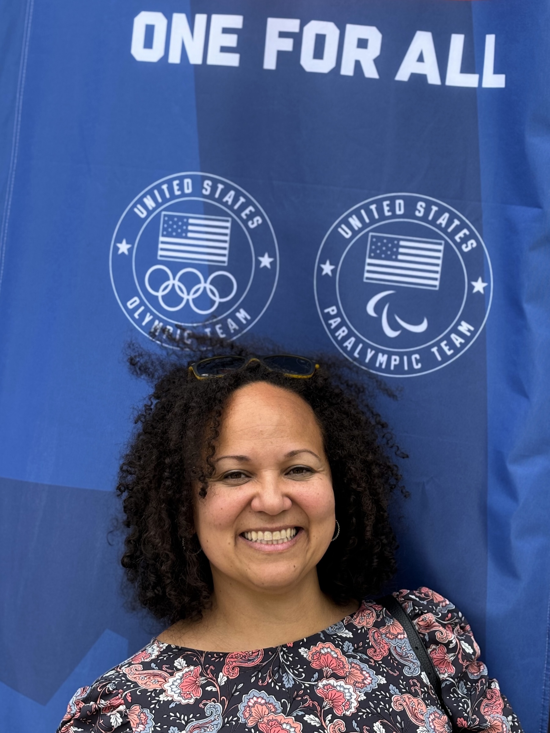 A woman smiles in front of a blue backdrop that says "ONE FOR ALL" above the United States Olympic and Paralympic team logos.