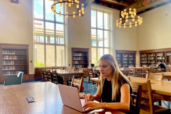 Young woman with blond hair sits at laptop in large ornate reading room