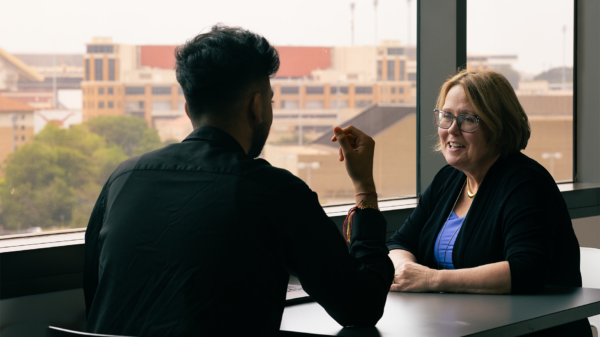 Two people sitting at a table talking with Darrell K Royal-Texas Memorial Stadium visible in the background through the window.