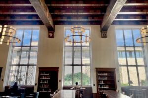 Students study at desks facing out the windows of The Hall of Texas, a high ceilinged library room with ornately painted beams, rafters and ceiling and circular chandeliers