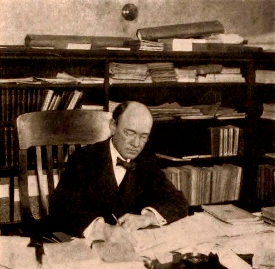 Black and white photo of bald man in bow tie writing at his desk