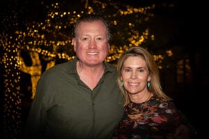 Married couple poses for a photo at night in front of holiday lights on trees.