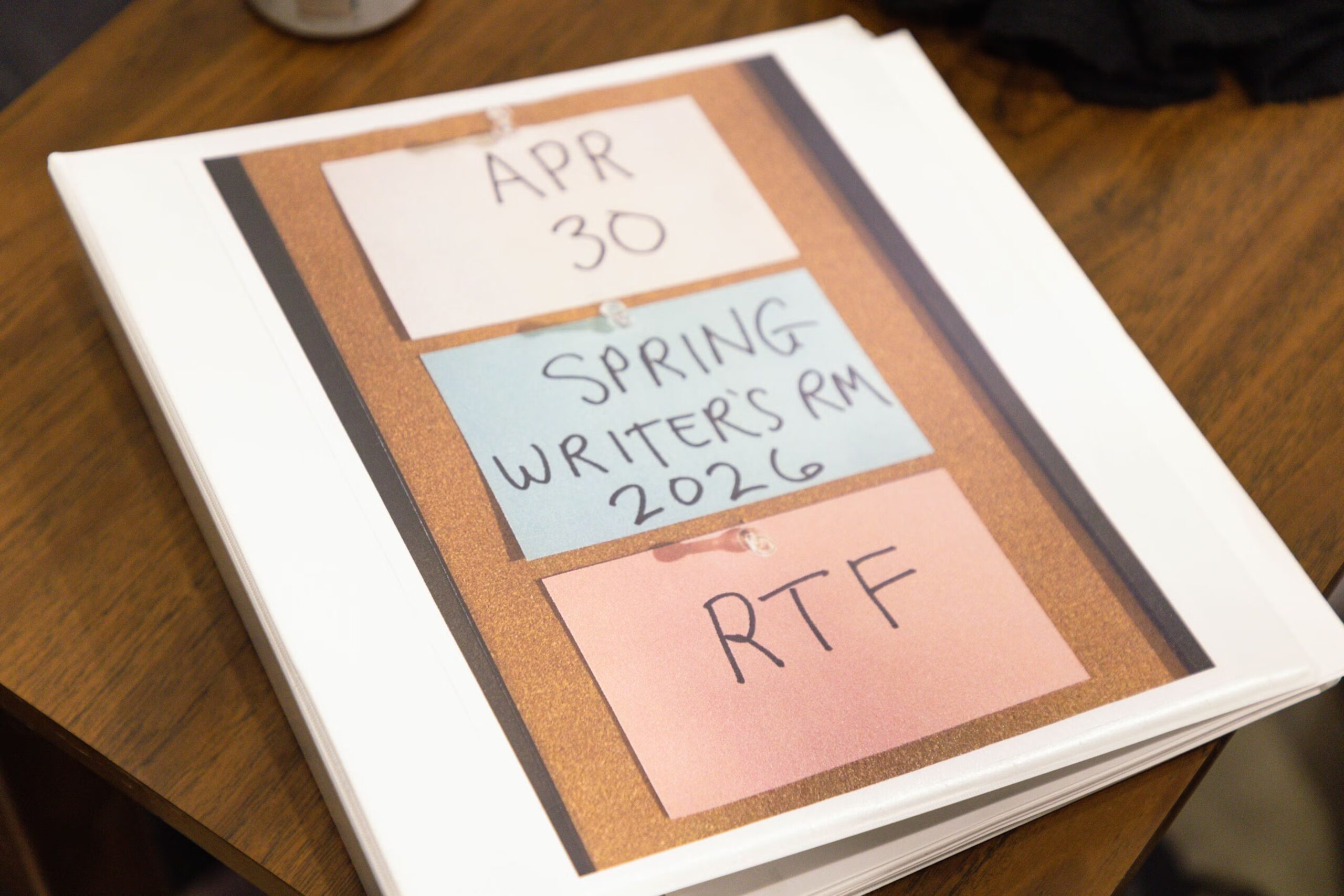 A photo of a bulletin board with three note cards that read "APR 30 / SPRING WRITERS' RM 2026 / RTF" in the cover of a white three-ring binder.
