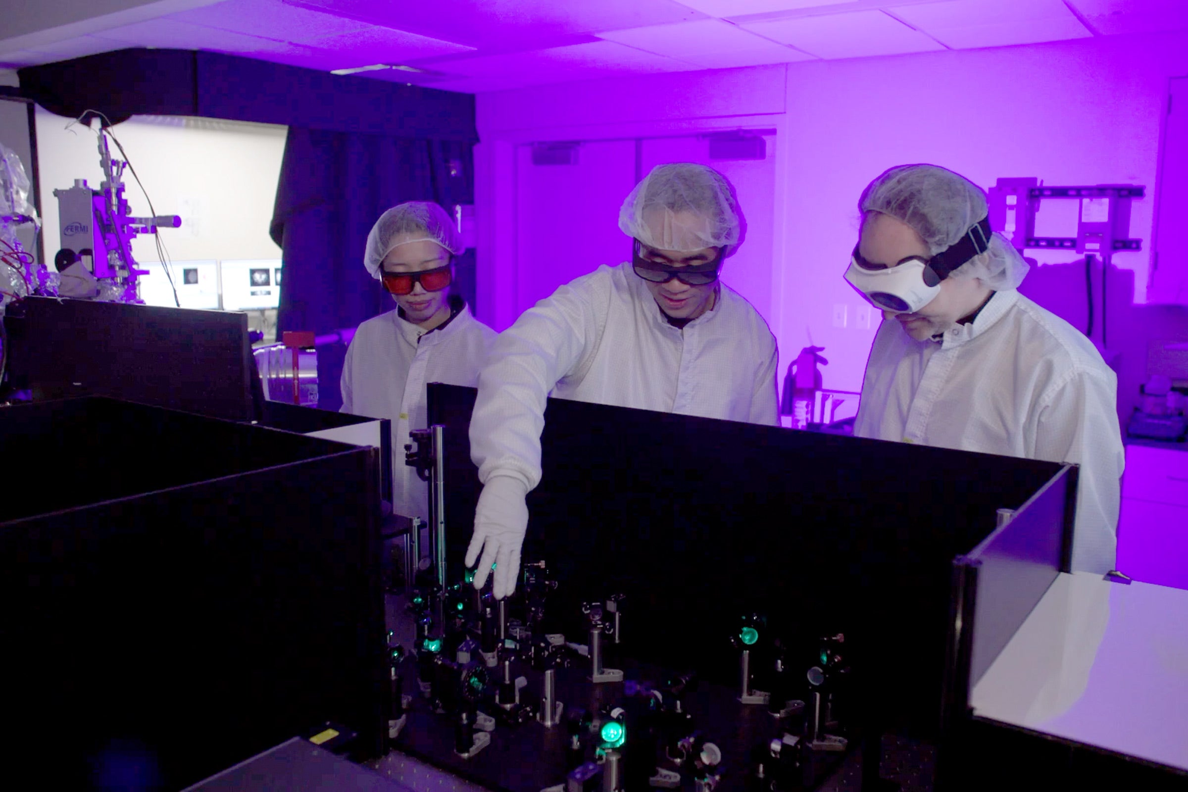 Three scientists wearing personal protective equipment work in a lab with purple lighting.