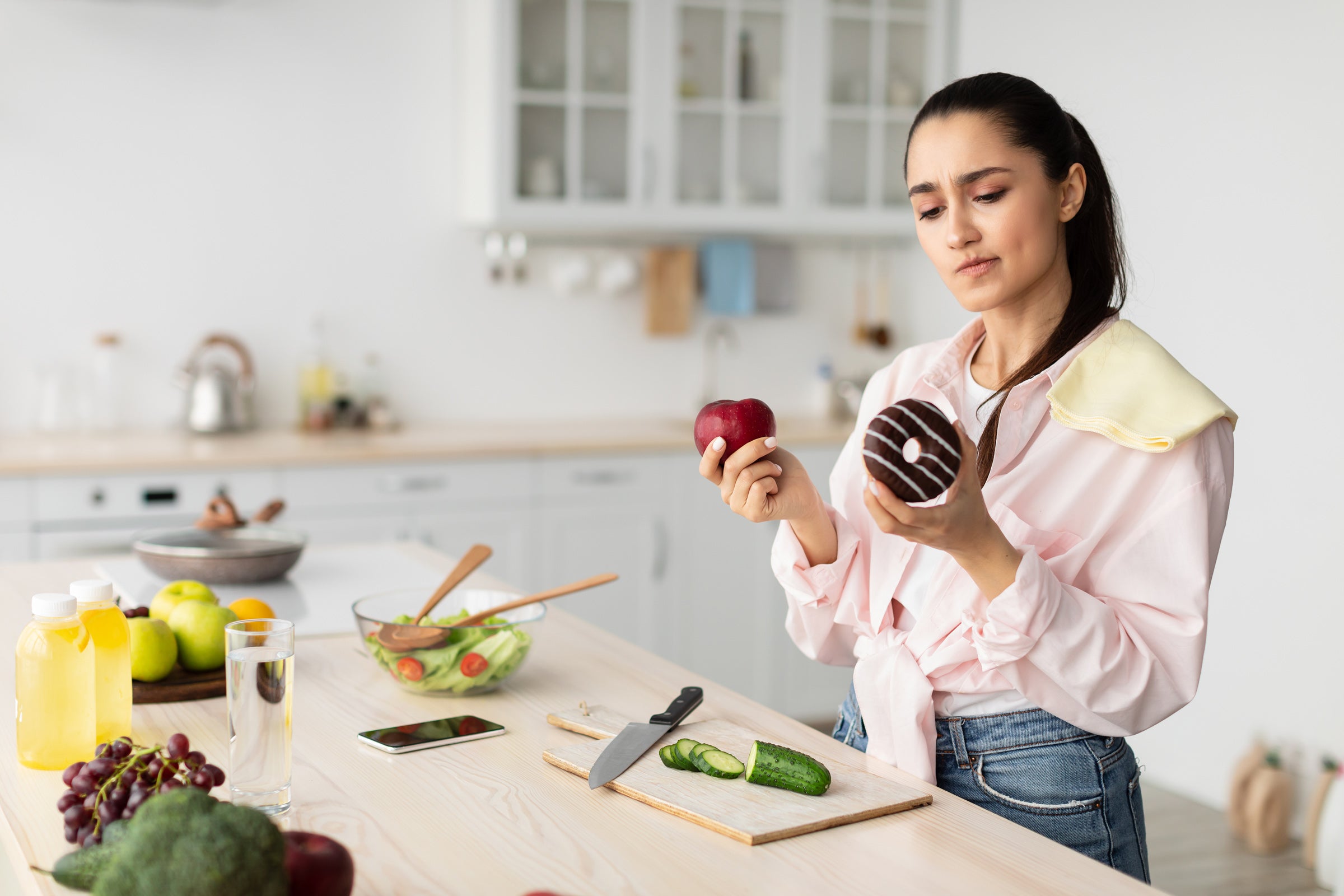 A woman in a pink shirt wears a conflicted expression on her face while holding an apple and a donut